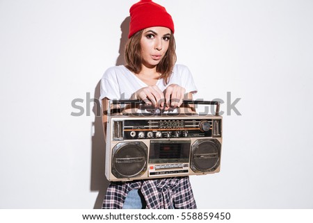 Picture of young pretty woman dressed in white t-shirt standing isolated over white background while holding tape recorder.