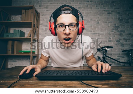 Young man playing game at home and streaming playthrough or walkthrough video. Funny teenager is excited because of seen virtual video. He is wearing red headphones and eyeglasses Royalty-Free Stock Photo #558909490