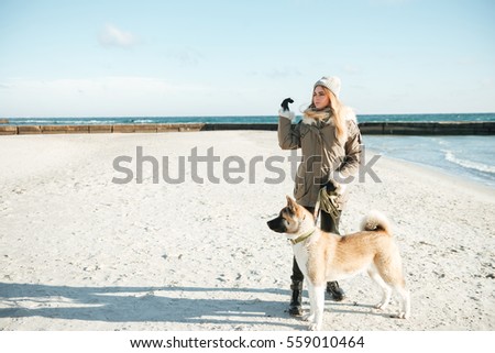 Picture of caucasian young woman walks in winter beach with dog on a leash.