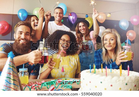 Group of six friends holding drinks at birthday celebration in room filled with colorful balloons