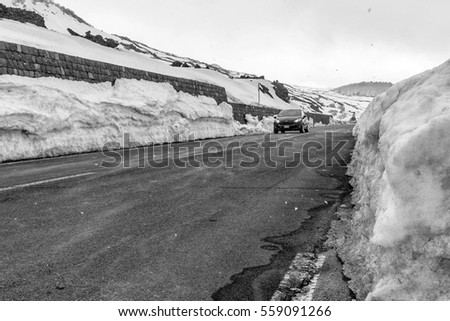 A small car runs along a stretch of road in the mountains while it snows and the edge of the road is covered by a high layer of snow, in black and white