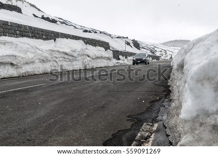 A small car runs along a stretch of road in the mountains while it snows and the edge of the road is covered by a high layer of snow