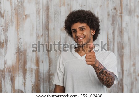 Positive curly haired man with beautiful tattoo showing thumbs up. While standing against old wooden background