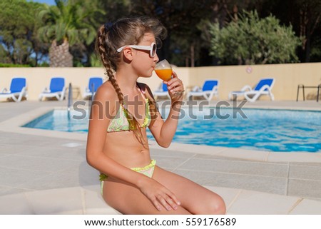 girl in bikini with glass of orange juice in hand sitting in swimming pool in hotel courtyard