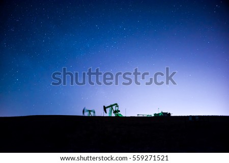 Two oil pump jack silhouettes backlit by city lights & stars