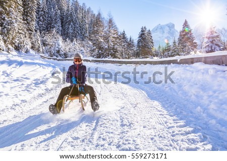 Cheerful girl riding a sled downhill on a snow covered sledge trail in a white sunny winter mountain landscape