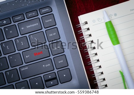 Laptop keyboard with visible red text "start", notebook and a pen, top view.