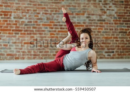 Portrait of attractive girl with bright red hair wearing gray and vinous sportswear. Model working out against brick wall, doing yoga or pilates exercise with mat on white wooden floor. Full length