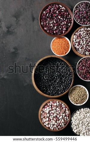 Set of various beans in bowls: white, black, purple and red speckled beans, red and green lentils. Black wooden background, top view.