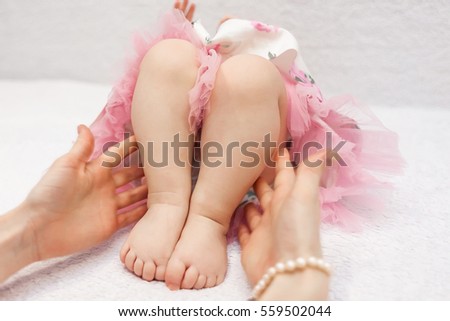 Mother holding her baby daughter's legs while baby girl lying on white soft blanket in cute pink dress
