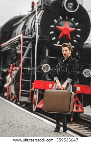A girl with a vintage hair and a suitcase stands near an old locomotive of a train