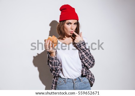 Picture of young hungry lady dressed in shirt in a cage print wearing hat standing isolated over white background and eating burger