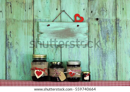 Blank mint green wood sign with red heart hanging on antique rustic wooden background over display of fruit jelly jars on shelf with red and white checkered fabric; holiday background with copy space