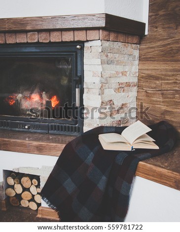 Black and blue blanket and open book which lies next to the stone fireplace and  firewoods. Royalty-Free Stock Photo #559781722