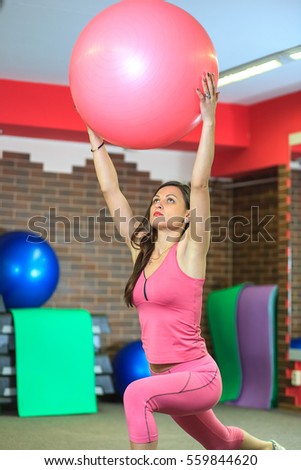 Young beautiful white girl in a pink sports suit does physical exercises with a fit ball at the fitness center. Hall fitness club for background. Interior of fitness club.