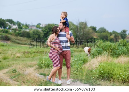 Happy pregnant family having fun in summer nature. A child on the shoulders of dad. Countryside, walk along rural road. Happy pregnant family kisses