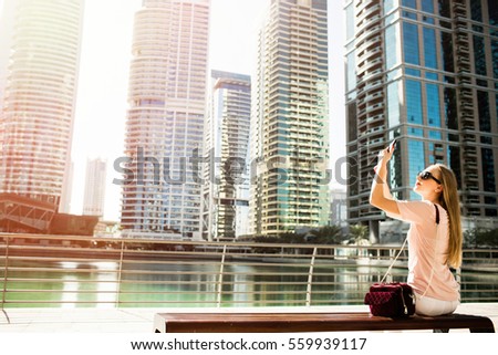 Girl takes picture on her phone sitting on the bench on embankment in Dubai