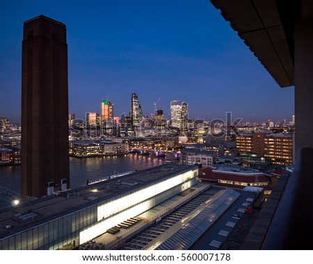 The cityscape and skyscrapers of The City of London at dusk, including the Heron Building, the Walkie-talkie and Tower 42 landmarks. The scene is framed by the Tate Modern chimney and rooftop.