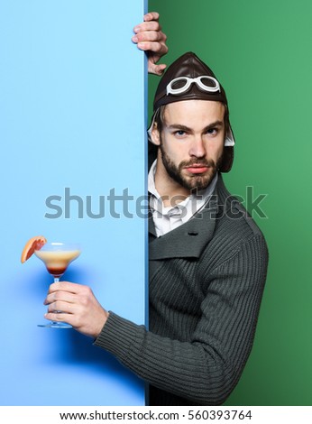 handsome bearded aviator man with long beard on serious face holding glass of alcoholic beverage in gray knitted sweater with hat and glasses on blue green studio background