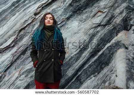 Portrait of a beautiful hipster girl on the background of the rocky cliffs. Dyed hair, blue, long. Kind and beautiful in a green coat.