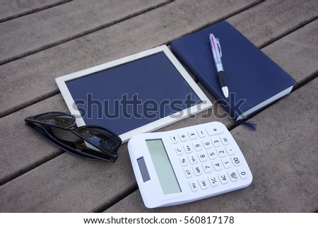 Small blackboard, white calculator, blue navy notebook and ball pen on wooden table.