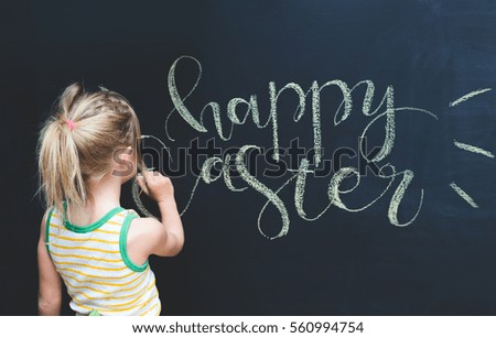 blond girl in preschool age writing Happy Easter with a chalk on the black board