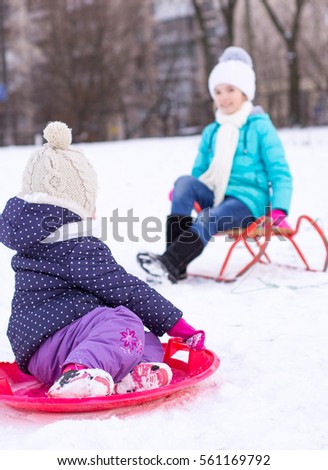 Sisters sledding on snow, all covered with snow on the walk in winter snowy forest or garden. Seasonal outdoor activities.