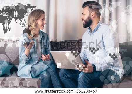 Bearded man and woman are sitting and discussing place to vacation. Girl uses smart phone, man holding laptop. Teamwork. In foreground semi-transparent virtual icons, image maps of world and stars.