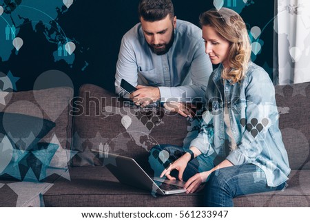 Young businesswoman sitting on sofa and using laptop.Nearby stands bearded businessman and looking on computer screen. Shopping online.In foreground semi-transparent virtual icons,image maps of world.
