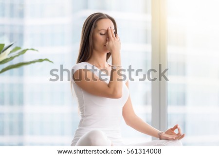Young attractive woman practicing yoga exercise, sitting in Sukhasana pose, performing Alternate Nostril Breathing technique, nadi shodhana pranayama, working out, wearing white sportswear, indoor  Royalty-Free Stock Photo #561314008