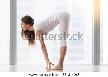 Young woman practicing yoga, standing in head to knees, Ardha uttanasana exercise, Half forward bend pose, working out, wearing sportswear, white t-shirt, pants, indoor full length, near floor window 