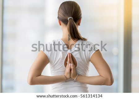 Young woman practicing yoga, making Namaste behind the back, working out, wearing sportswear, white t-shirt, indoor, against window with city skyscraper, rear view 