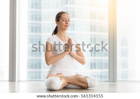 Young attractive woman practicing yoga, sitting in Padmasana, exercise, Lotus pose, namaste, working out, wearing sportswear, white t-shirt, pants, indoor full length, near floor window with city view Royalty-Free Stock Photo #561314155