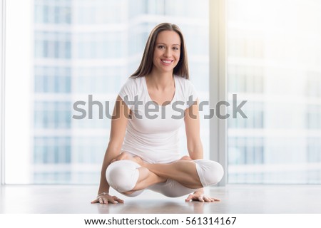 Portrait of young smiling woman practicing yoga, standing in Scale exercise, Tolasana pose, working out, wearing white sportswear, full length, near floor window with city view, looking at camera 