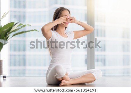 Young woman practicing yoga, sitting in Sukhasana pose, performing Humming Bee Breathing exercise, Bhramari Pranayama technique, working out, wearing sportswear, white t-shirt, pants, full length  Royalty-Free Stock Photo #561314191