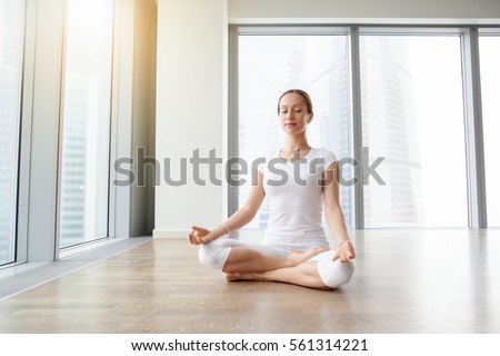 Young attractive woman practicing yoga, sitting in Ardha Padmasana exercise, Half Lotus pose, working out, wearing white t-shirt, pants, meditation session at floor window with city view. Full length  Royalty-Free Stock Photo #561314221