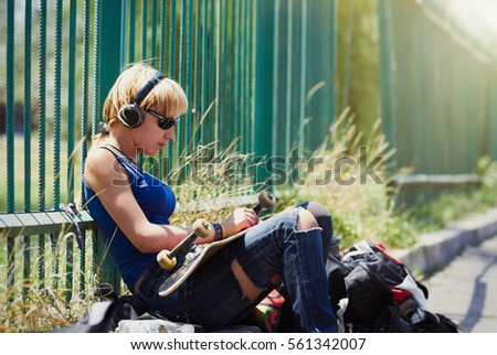 Young skater girl listening to music in wireless headphones in skatepark outdoor. White skateboarded female athlete chilling in skate park in sunny summer day. Teenager punk rock chick in torn jeans 
