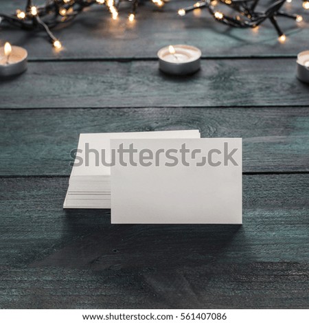 A photo of a blank white thick cardboard business card next to a pile of them, on a dark wooden texture with candles in the background. A square mockup or a banner with copyspace