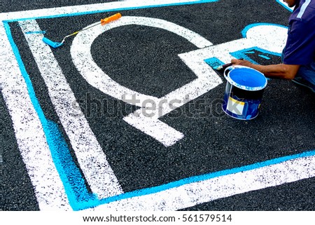 Close up of man painting empty handicapped reserved parking space with wheelchair symbol on black asphalt. No parking white painted letters and blue diagonal lines in background.