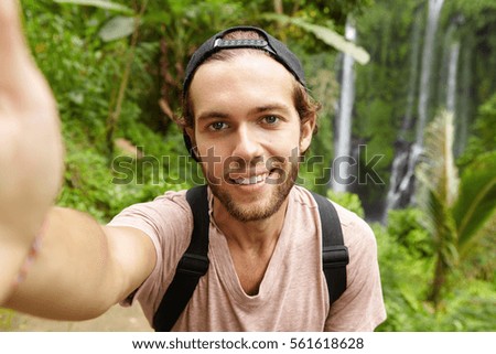 Close up view of handsome Caucasian hiker wearing snapback looking at camera with happy smile while taking selfie with amazing landscape with waterfall in background. Travel, adventure and tourism