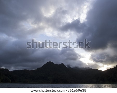 Cloudy sunset over mountain lake. Spectacular stormy clouds under sunlight. Evening scene of wild nature. Black silhouette of old mountains surrounding volcanic lake. Evening skyscape view photo