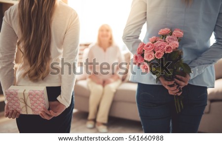 Teenage girl and her mom are hiding flowers and a gift box for their beautiful granny behind backs while grandma is sitting on couch at home Royalty-Free Stock Photo #561660988