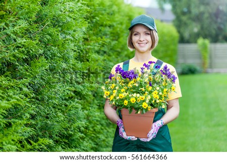professional gardener at work. smiling young woman gardener holding pot with flowers in the yard. garden worker planting flowers. gardening service and business concept