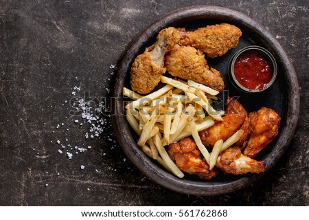 Fast food fried crispy and spicy chicken legs, wings and french fries potatoes with salt and ketchup sauce served in stone plate over dark texture background. Top view, space for text Royalty-Free Stock Photo #561762868