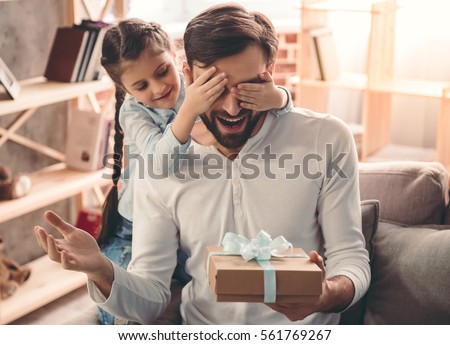 Cute little girl is giving her handsome father a gift box. Both are sitting on couch at home and smiling Royalty-Free Stock Photo #561769267