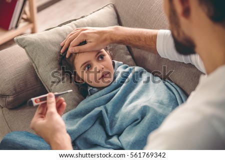Sick little girl covered in blanket is lying on couch while her father is taking her temperature Royalty-Free Stock Photo #561769342