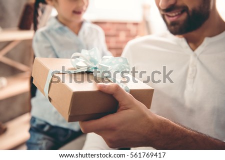 Cute little girl is giving her handsome father a gift box. Both are sitting on couch at home and smiling