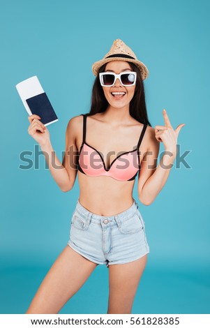 Picture of happy young lady with long hair wearing hat and dressed in swimwear posing over blue background holding passport