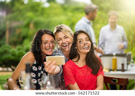 Group of female friends in their thirties taking a selfie during a party on a terrace. In the background a beautiful garden where their men doing a bbq. Shot with flare