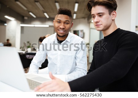 Photo of two students sitting in library and using laptop computer. African guy looking at camera. Caucasian man look at laptop.